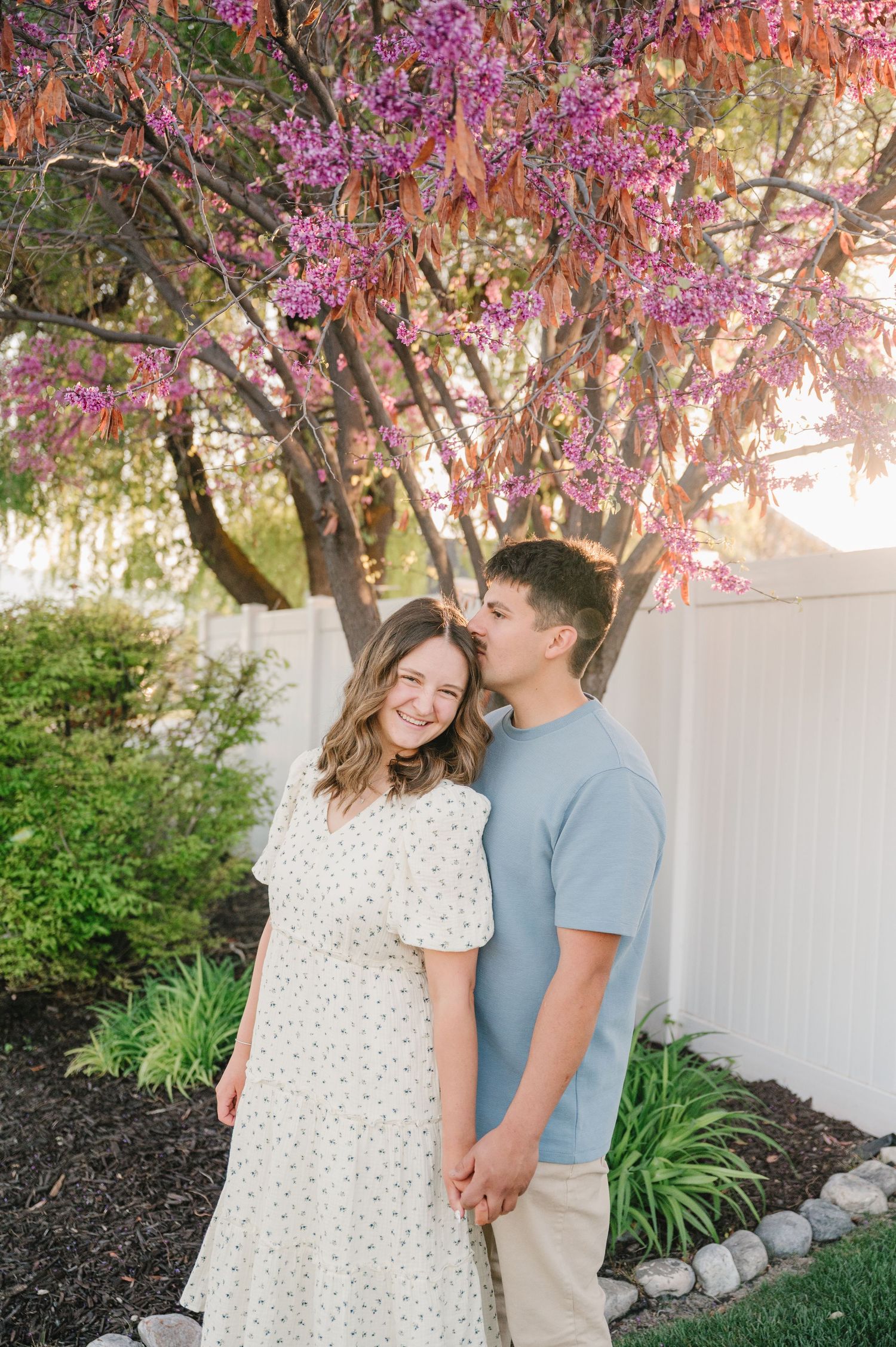 A couple shares a romantic moment under a blooming spring tree with pink blossoms against a white fence.