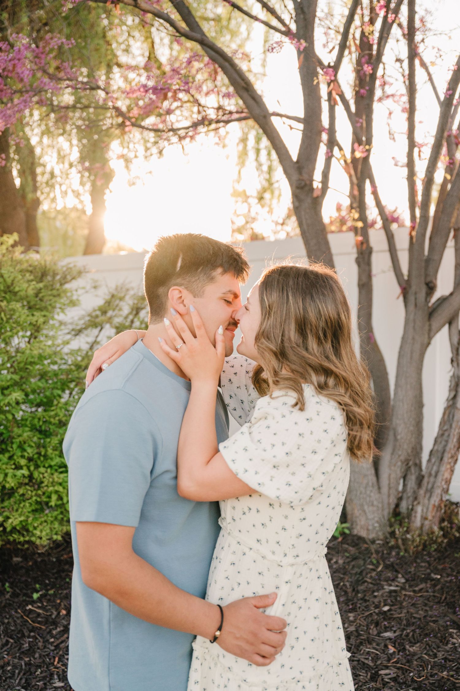 A romantic couple shares a tender embrace under flowering trees at sunset in a garden setting.
