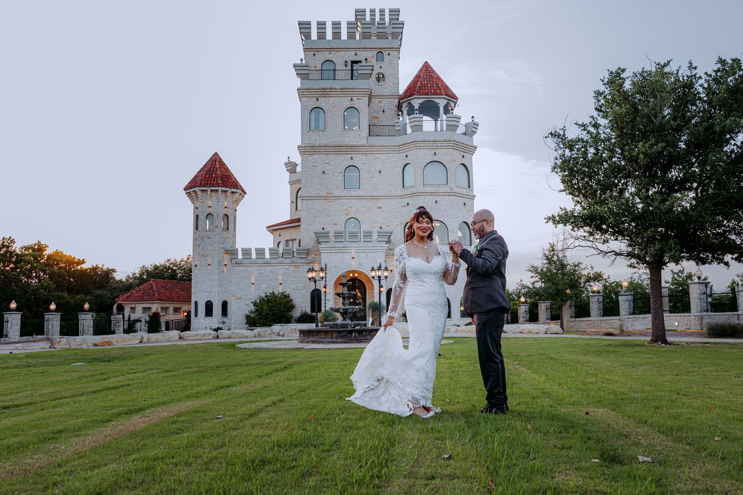 A bride and groom dance on a manicured lawn in front of a white fairytale castle with red-topped turrets.