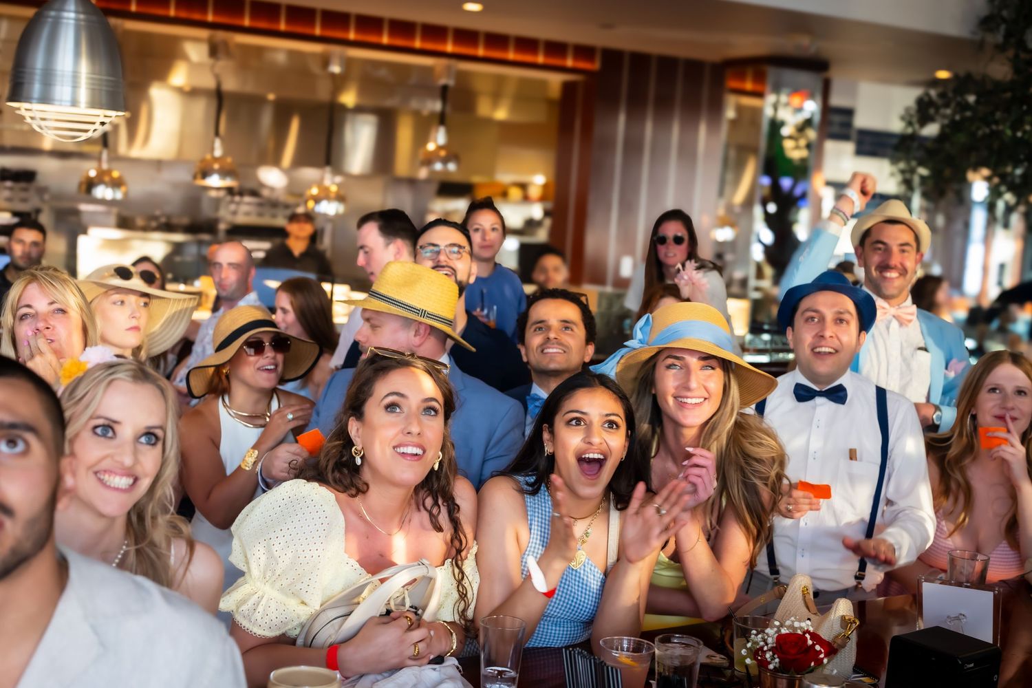 Excited wedding guests react with joyful expressions and wide smiles while watching a celebratory moment at a reception.