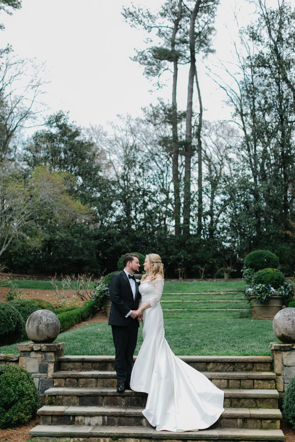 A couple poses romantically on stone garden steps surrounded by tall trees and landscaping during their wedding day.