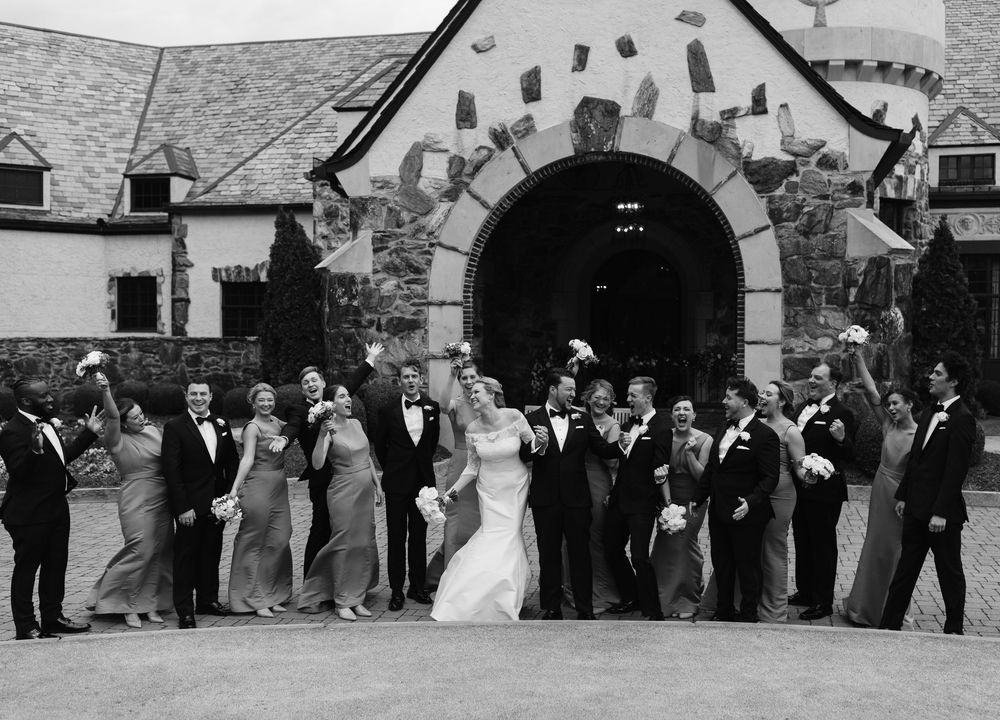 A wedding party poses in a curved line formation in front of a stone archway entrance at a historic venue.