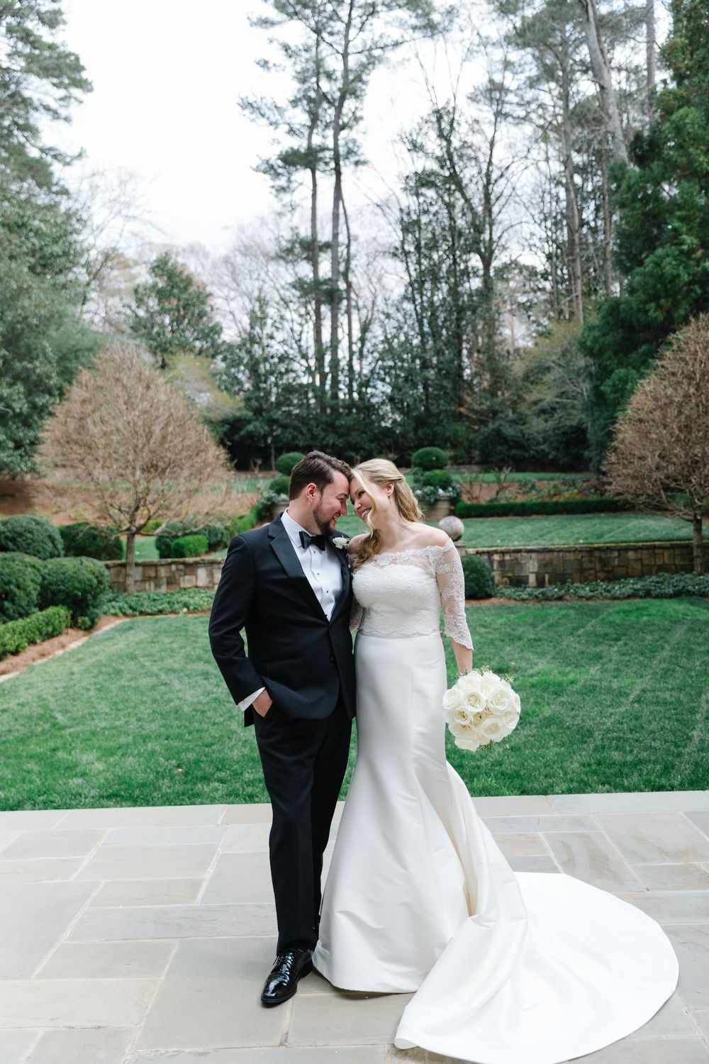 A couple shares an intimate moment in a formal garden setting with stone patio and manicured hedges.