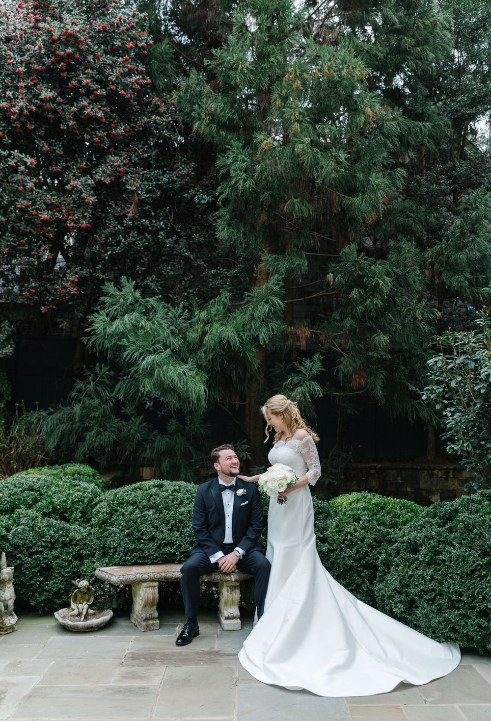 A couple in wedding attire poses in a garden setting with lush greenery and manicured boxwood hedges.