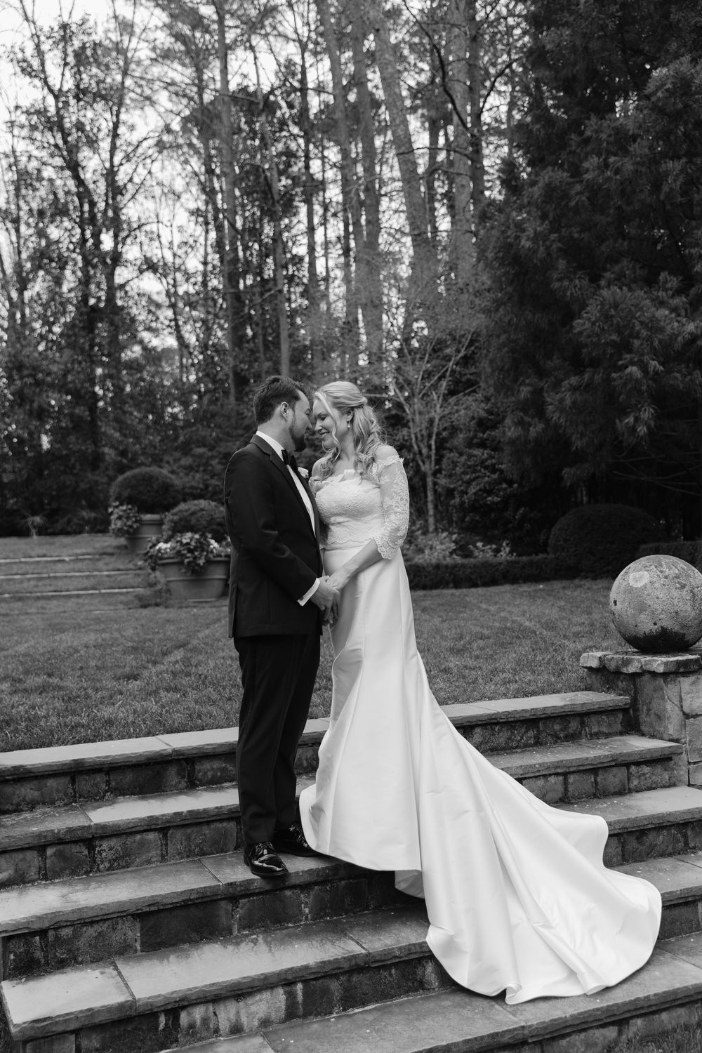 A romantic black and white wedding photograph capturing a couple on stone steps surrounded by tall trees.