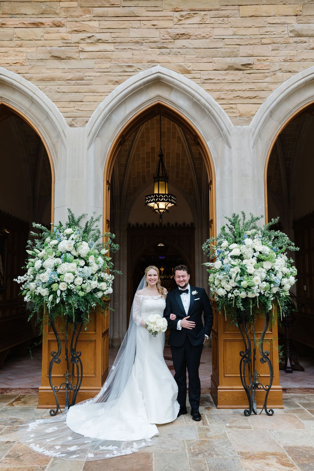 Cathedral entrance decorated with two large white floral arrangements and stone archways for an elegant wedding ceremony.