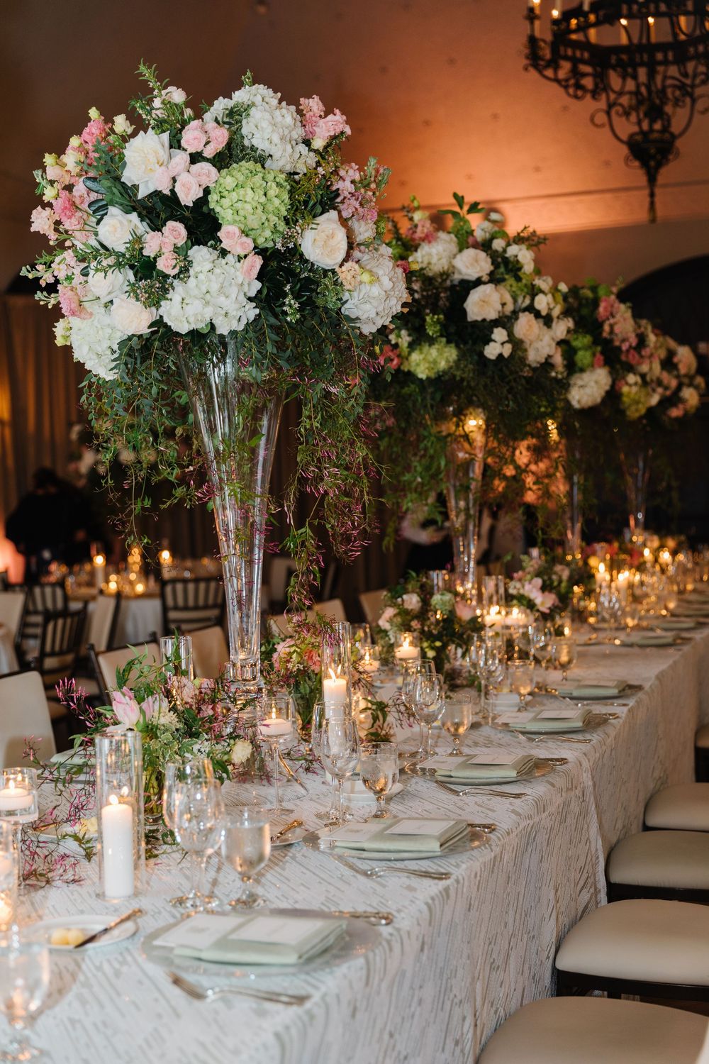 Elegant wedding reception table with tall floral centerpieces, white lace tablecloth, and glowing candlelight in warm ambiance.