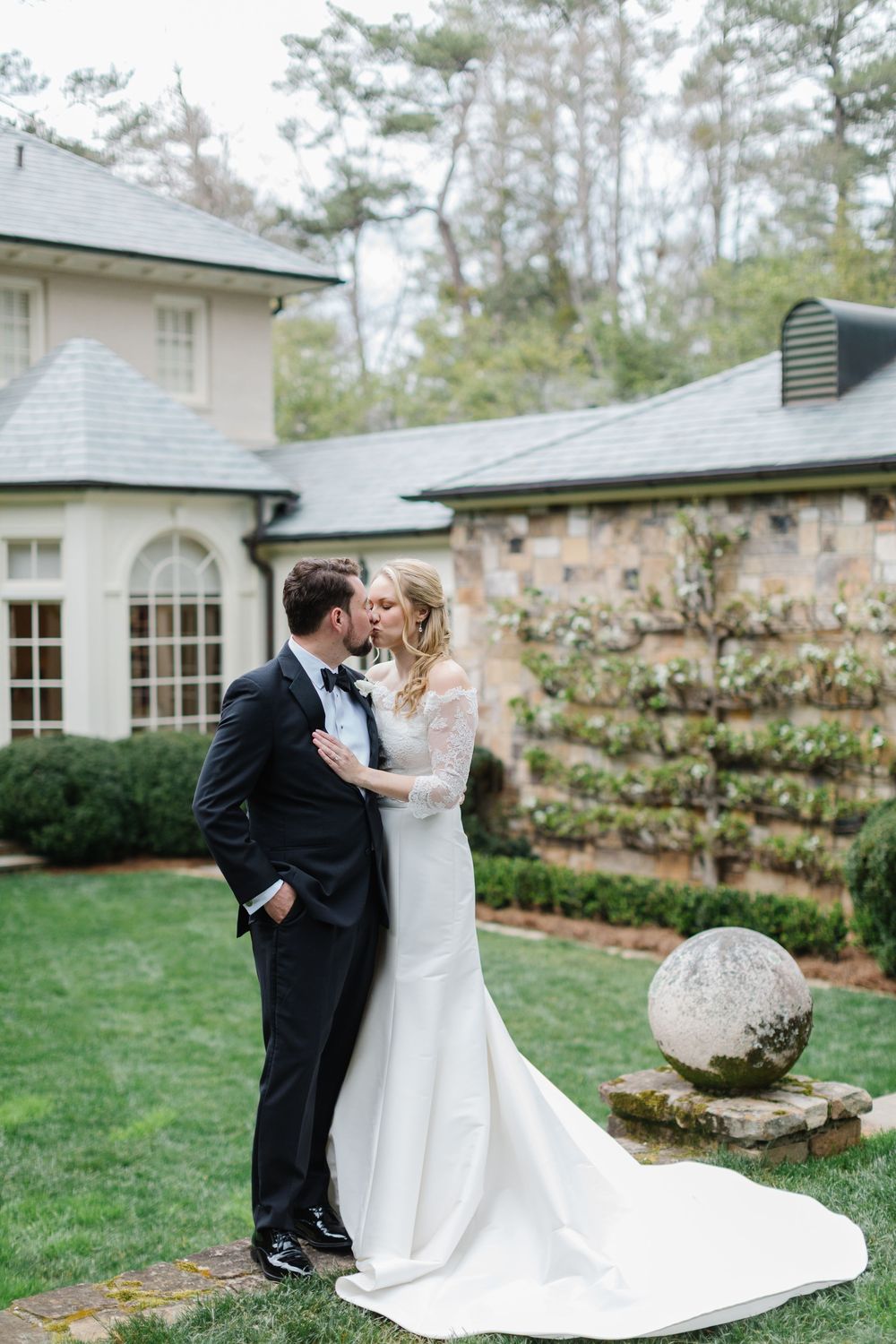 Elegant wedding couple embraces on manicured lawn in front of stone mansion with trailing vines and arched windows.