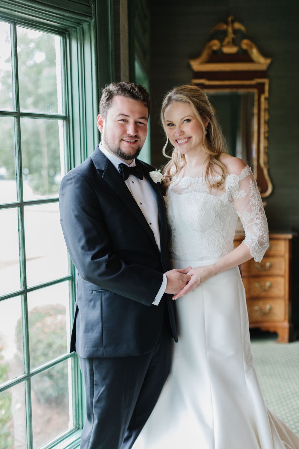Elegant couple poses by bright window in formal wedding attire featuring lace sleeve gown and black tuxedo.