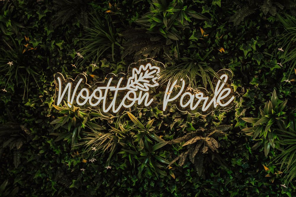 An illuminated 'Wootton Park' neon sign glows against lush green foliage.