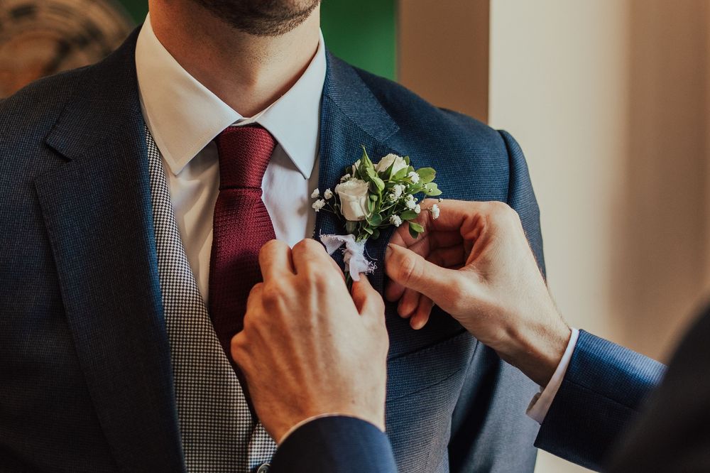 Groom's boutonniere being pinned to navy suit jacket during wedding preparations.