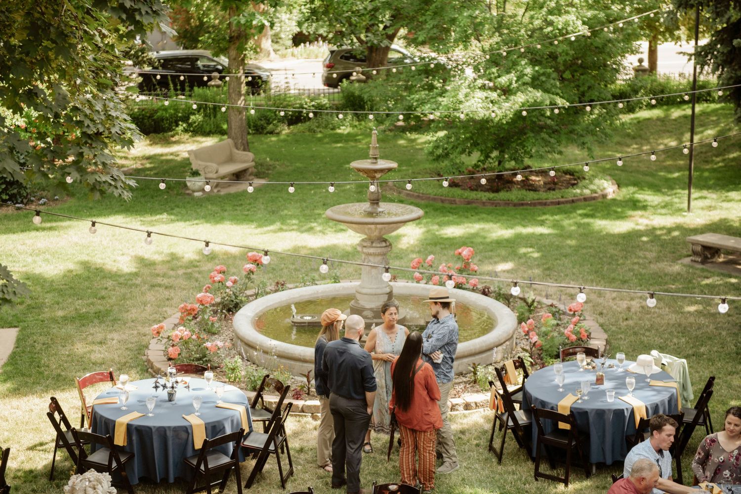 Elegant outdoor garden reception features round tables and chairs arranged around a decorative tiered fountain.