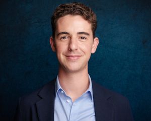 Professional headshot photo showing a young person in a navy suit jacket and light blue dress shirt against a textured blue background.