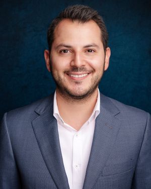 Professional corporate portrait of a person in a gray suit and white shirt against a blue textured backdrop.