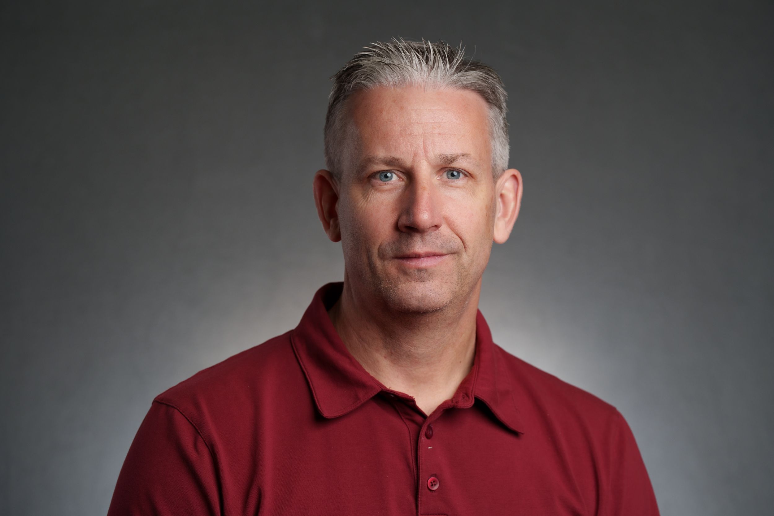 Professional headshot portrait of someone in a red polo shirt against a gray studio background.