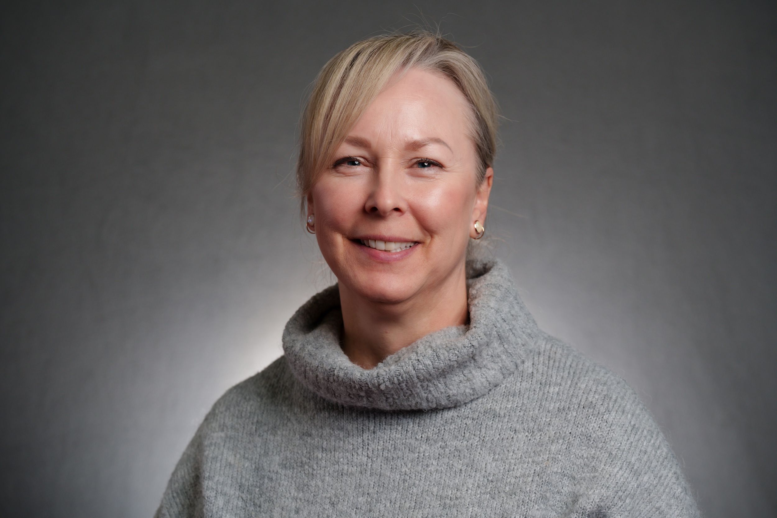 Professional portrait of someone wearing a cozy grey turtleneck sweater against a dark gradient background.