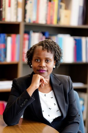Professional in a black blazer and patterned white blouse sits thoughtfully at desk with library bookshelves in background.