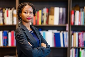 Professional in dark blazer stands confidently with arms crossed in front of library bookshelf.