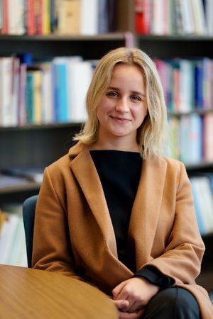 Professional in a brown coat sits at wooden table with bookshelves in background.