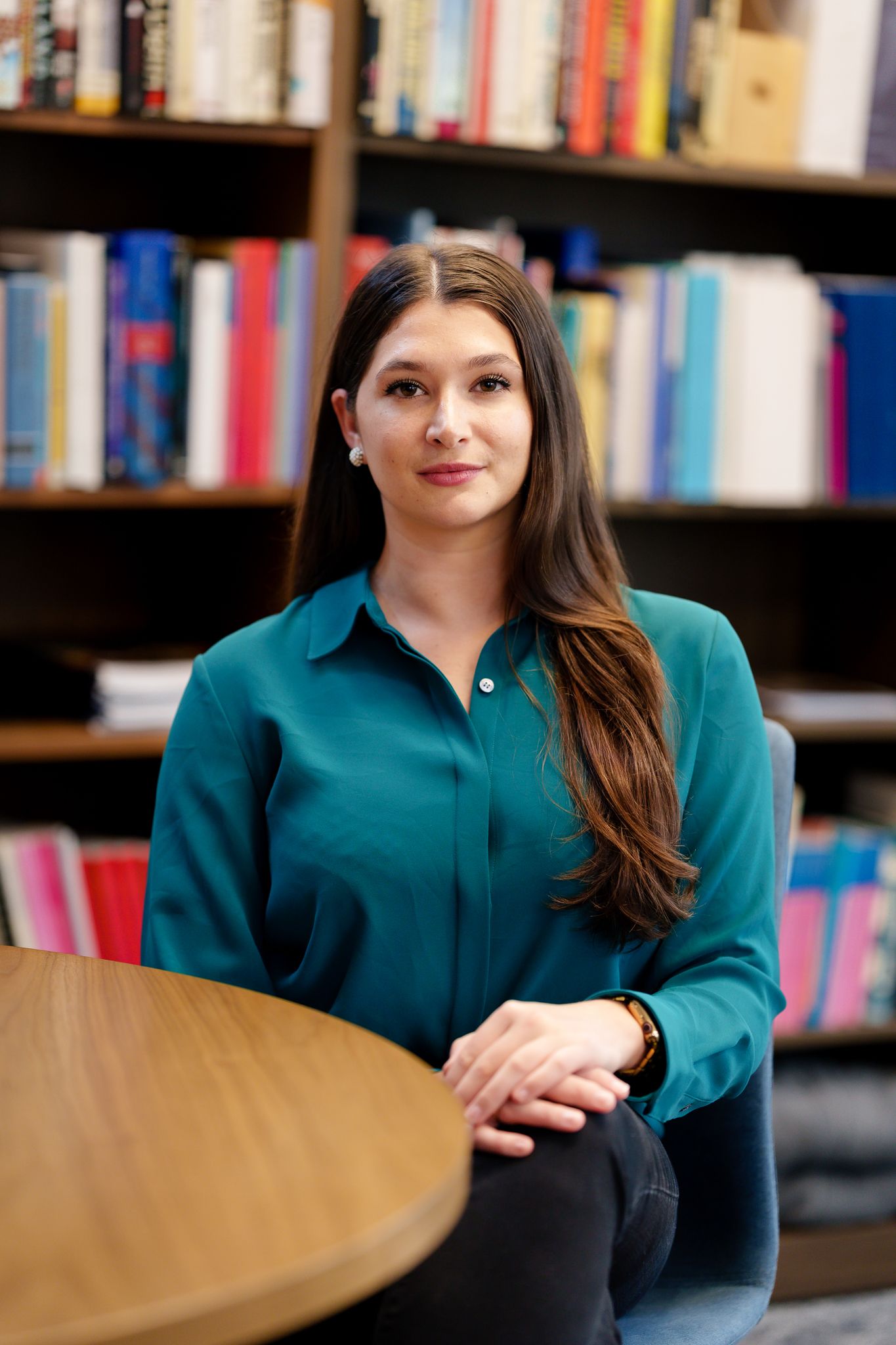 Professional in a teal blouse sits at a round wooden table with bookshelves in background of library or office setting.