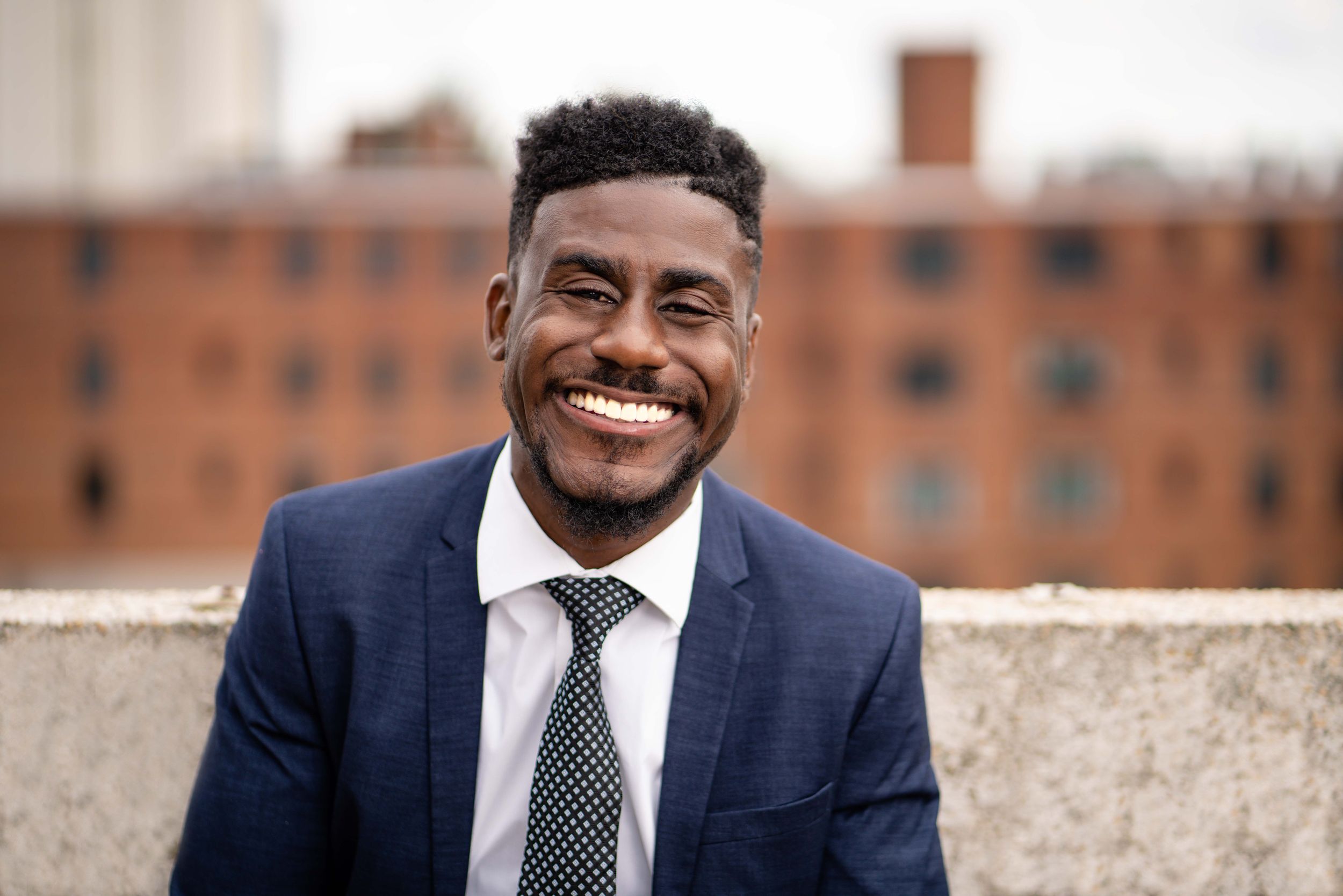 Professional wearing a navy blue suit and striped tie smiles warmly against a brick building background.