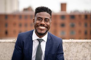 Professional wearing a navy blue suit and striped tie smiles warmly against a brick building background.