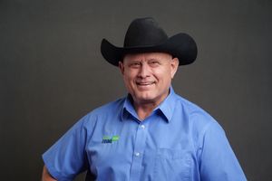 A person in a light blue western-style shirt and black cowboy hat smiles against a dark background.