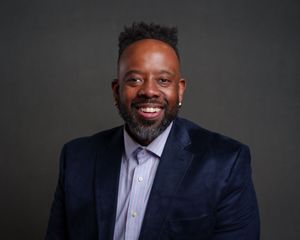 Professional headshot of someone wearing a navy velvet blazer and lavender dress shirt against a dark background.