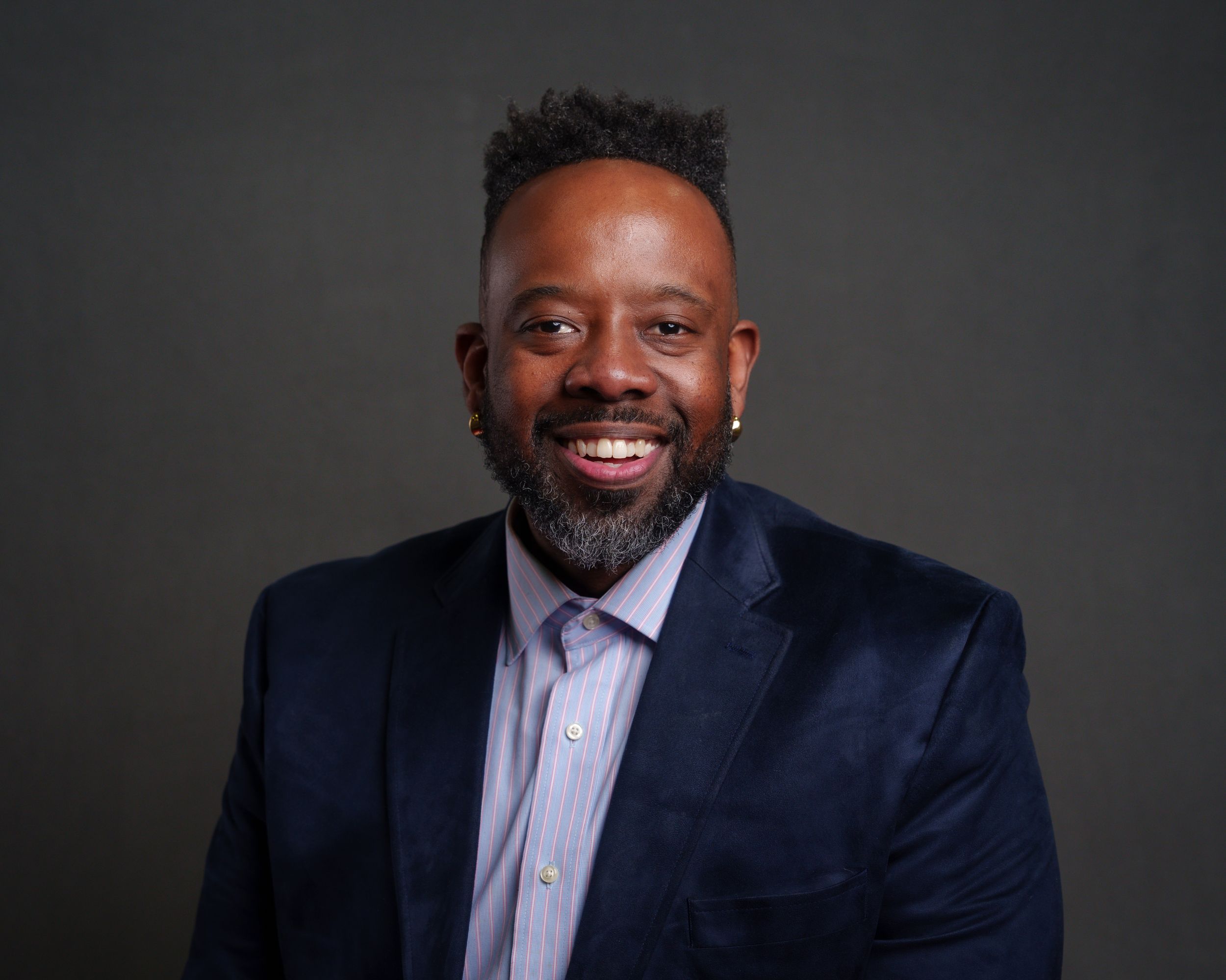 Professional headshot of someone wearing a navy velvet blazer and lavender dress shirt against a dark background.