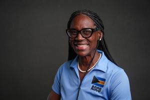 Professional portrait of educator wearing glasses and blue NSU polo shirt smiling warmly against dark background.