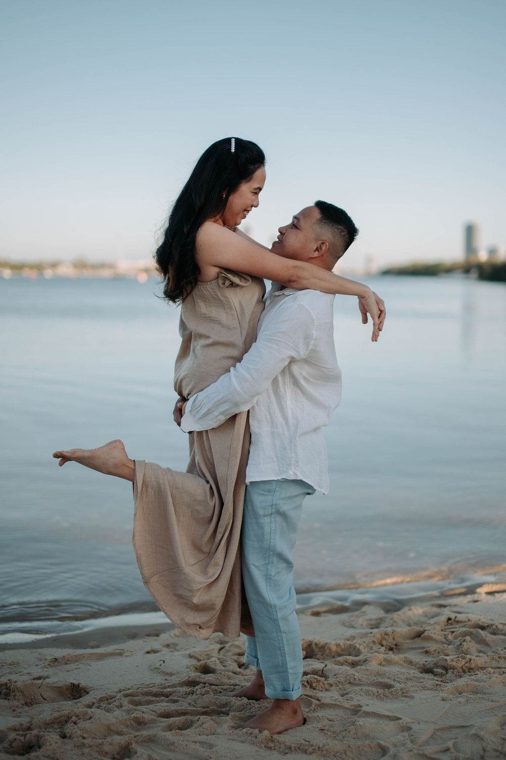 A couple embraces romantically on a beach at sunset with calm water and cityscape in the background.