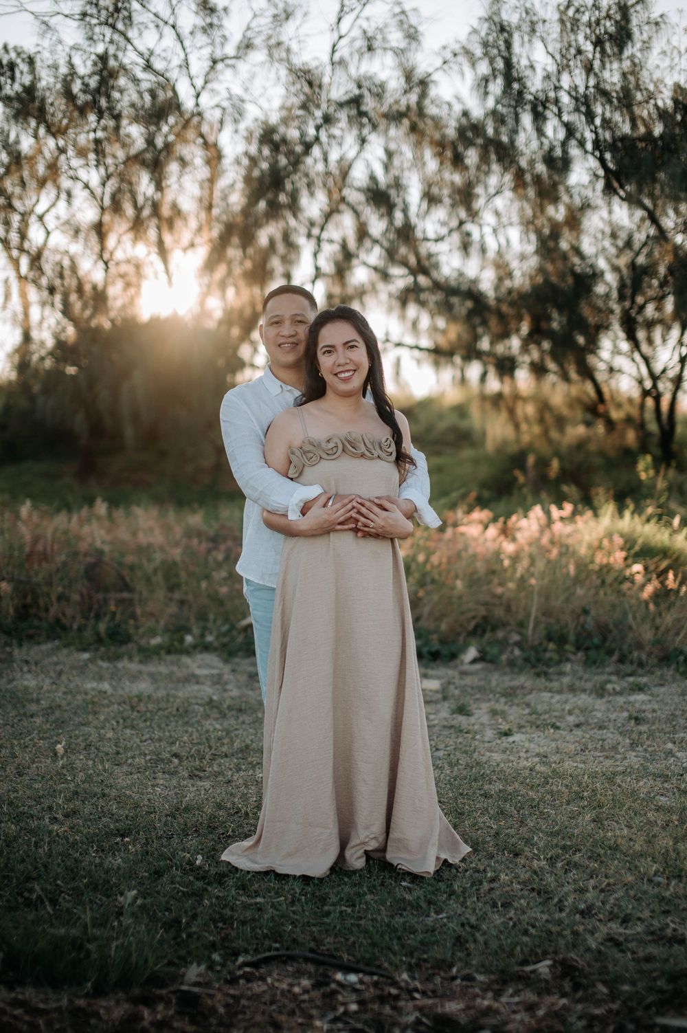 A romantic couple embraces in a misty sunset setting under tall trees casting long shadows in a field.