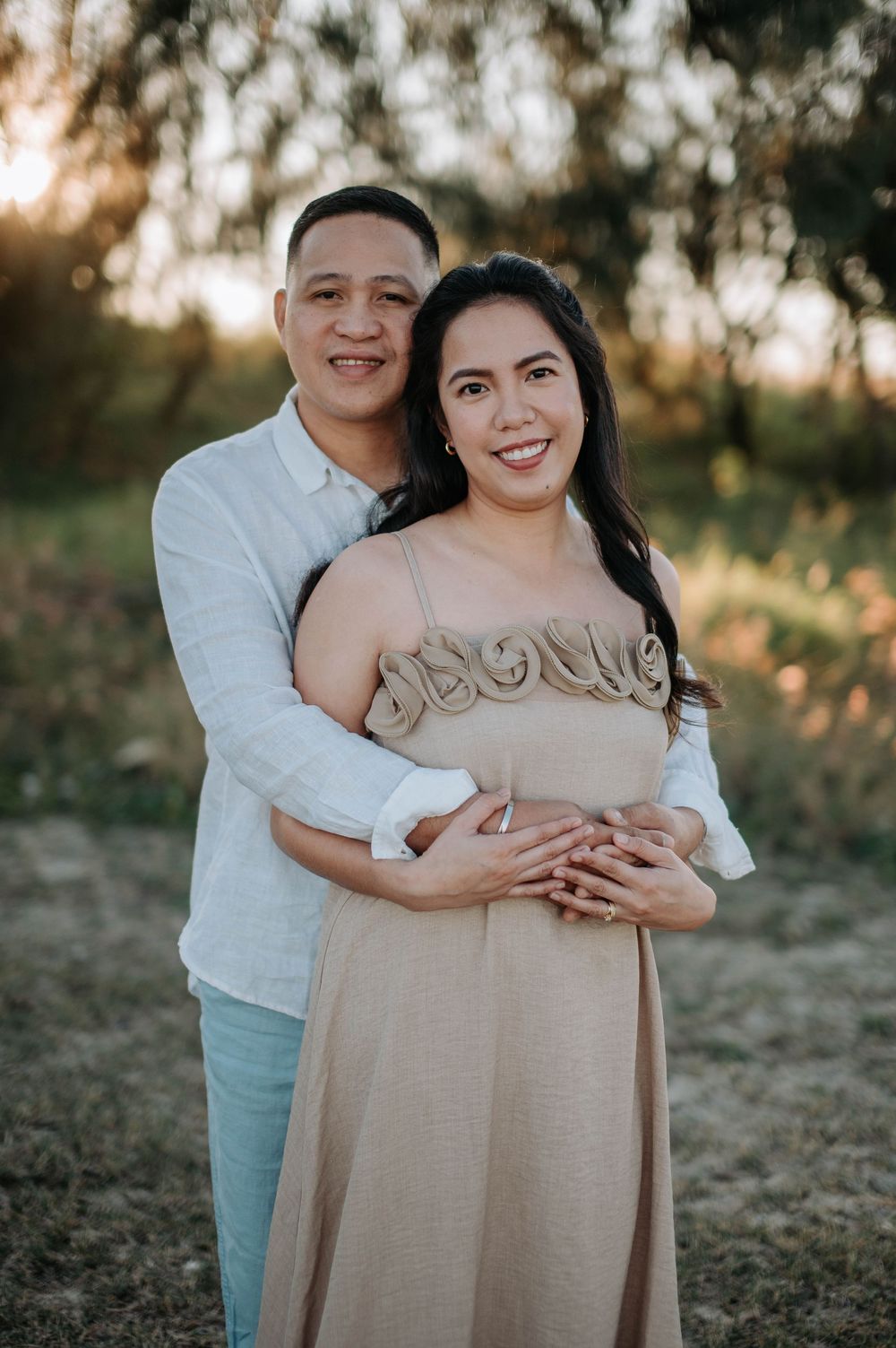 A romantic couple embraces in a golden sunset setting against trees during an outdoor portrait session.