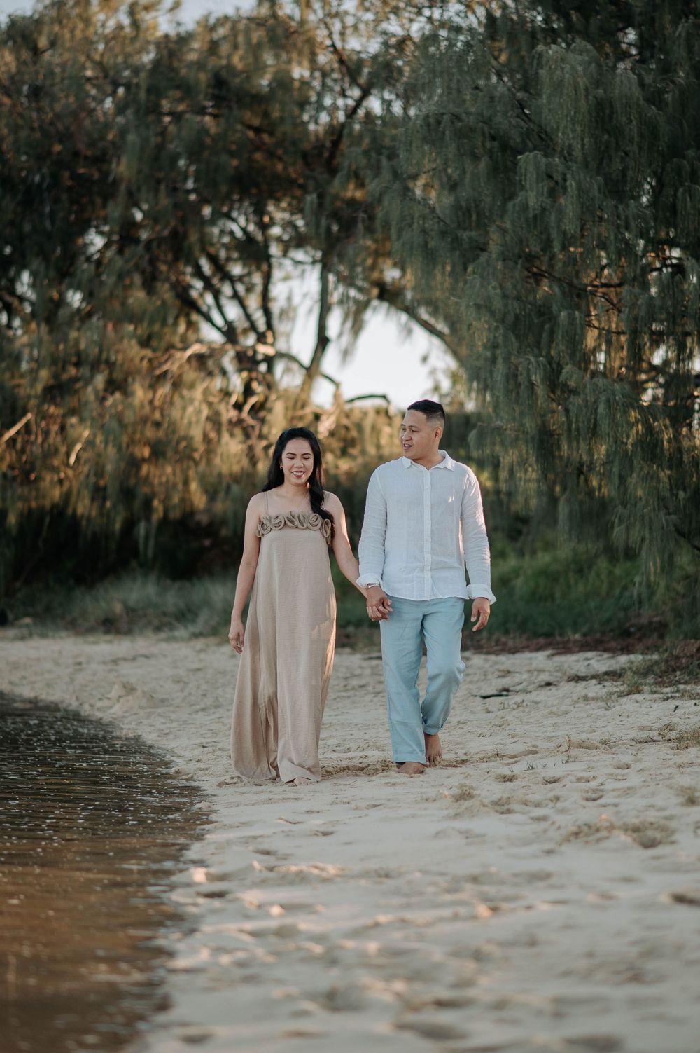 A couple strolls together along a scenic beach path with willow trees and golden sunlight in the background.