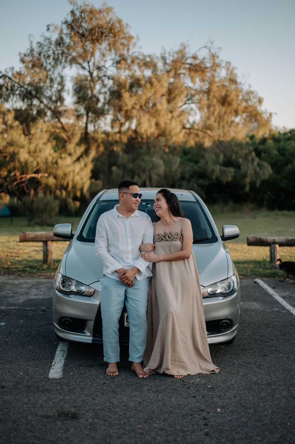 Couple in formal attire poses together in front of silver car during golden hour with trees in background.