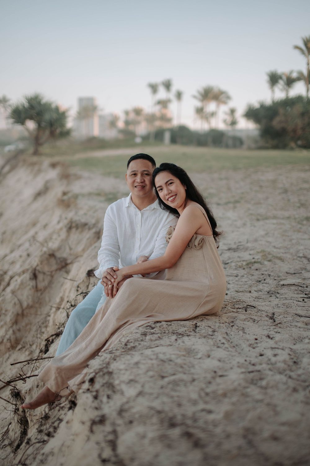A couple sits together on sandy terrain at sunset with palm trees and buildings visible in the background.