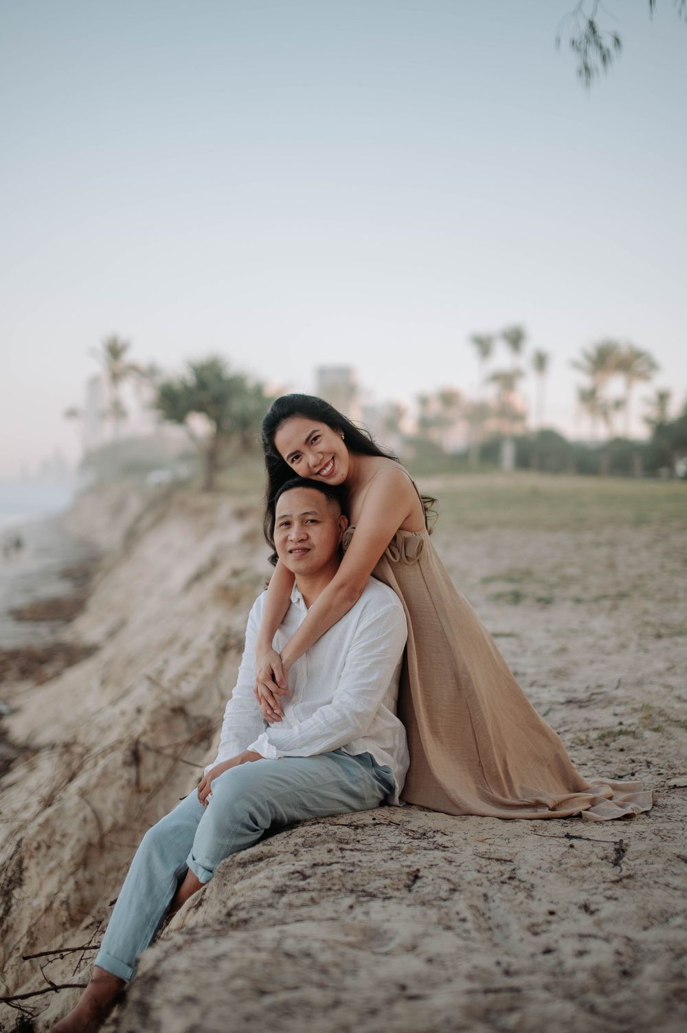 A couple embraces on a hilltop during a romantic photo session with palm trees and coastline in the background.
