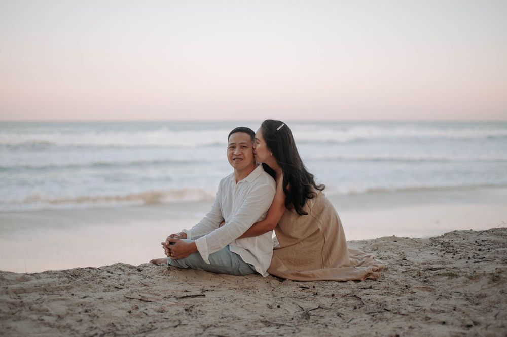 A couple sits together on the sandy beach at sunset, with gentle waves rolling in the background.
