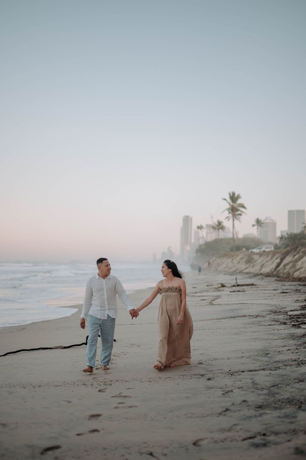 A couple walks hand in hand along a misty beach at sunset with palm trees and buildings visible in the background.