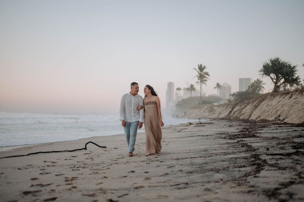 A couple walks along a misty beach at sunset with palm trees and cliffs visible in the background.