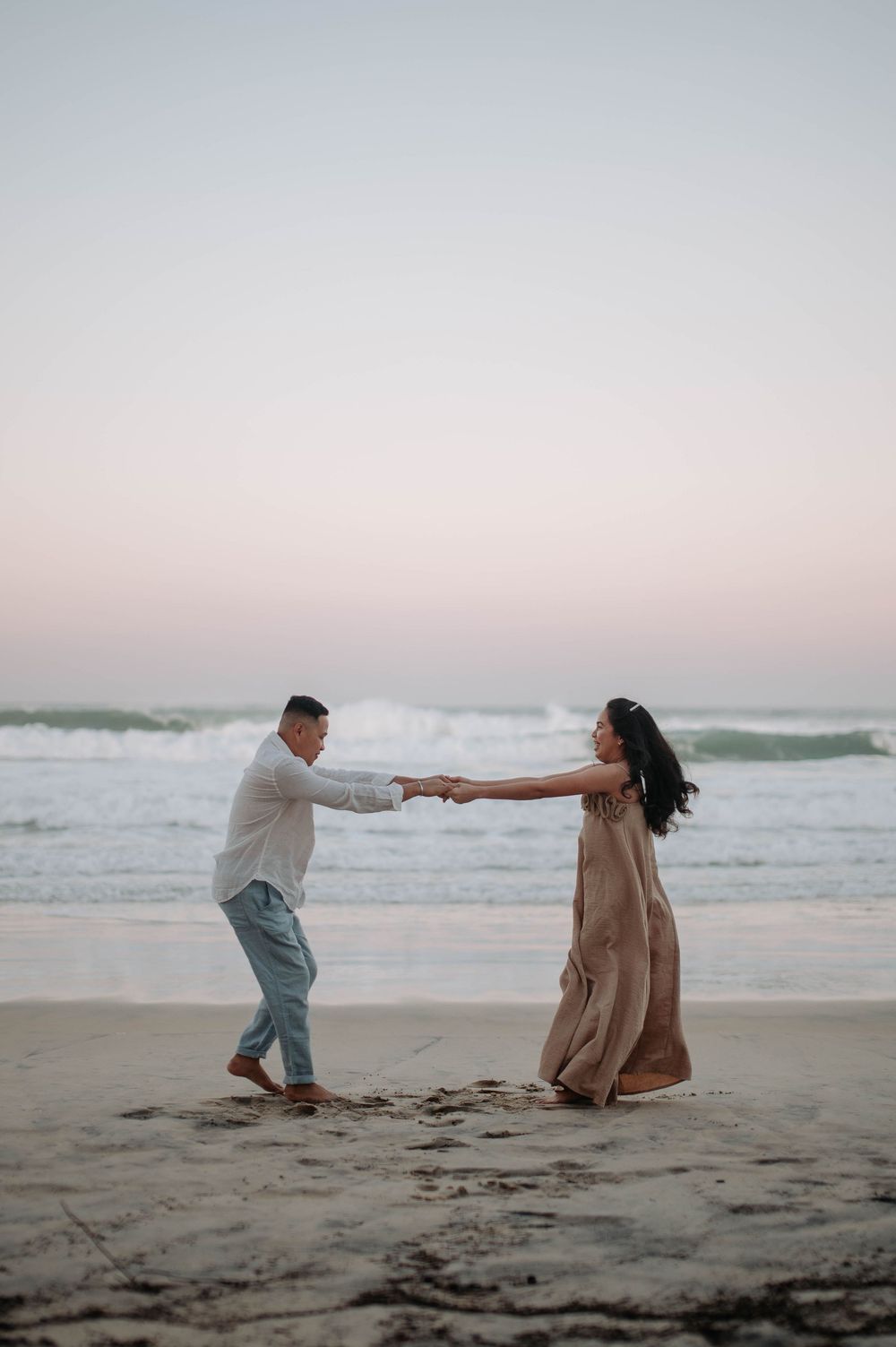 A couple dances joyfully together on a sandy beach at sunset with crashing waves in the background.