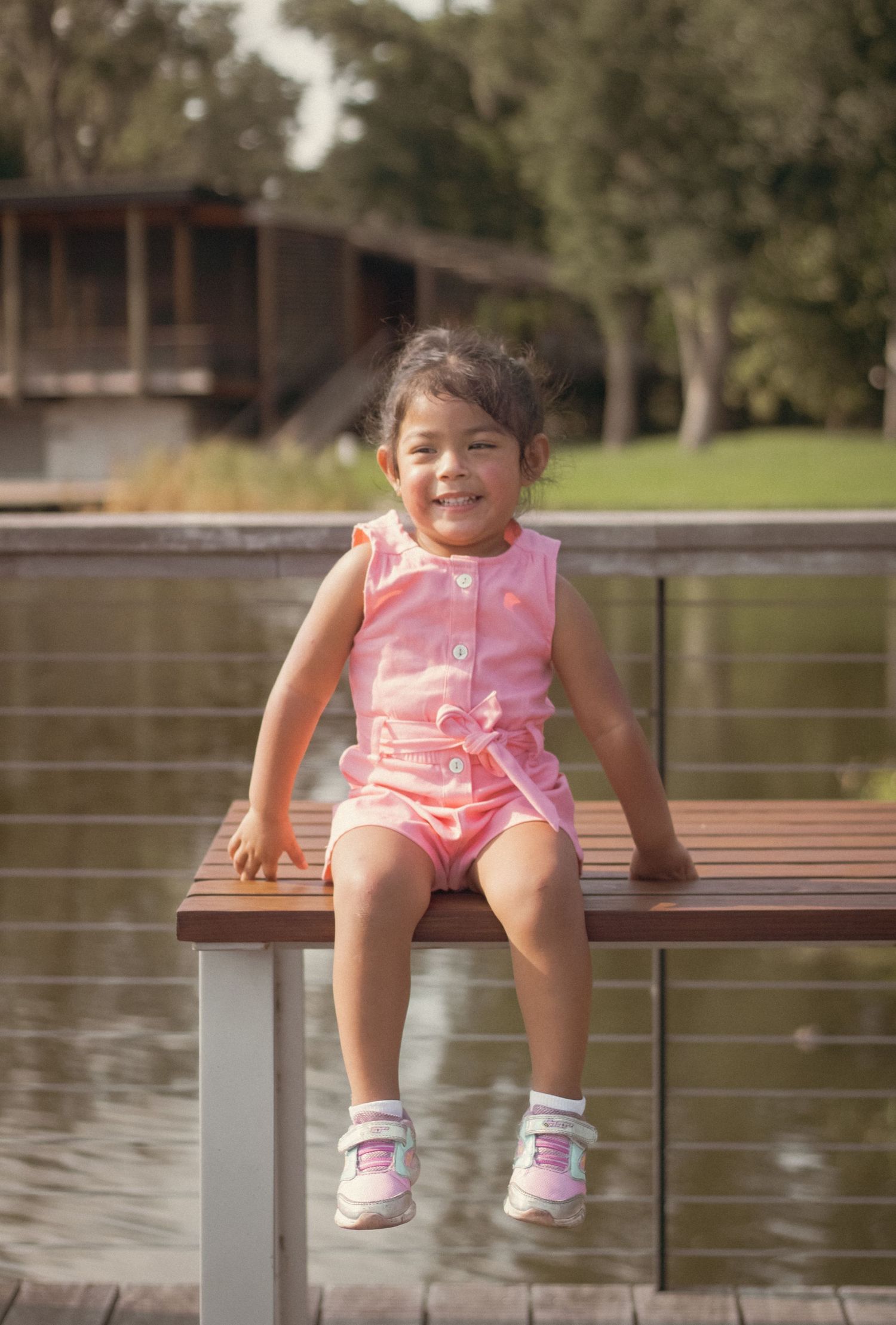Happy little girl in pink outfit sitting on a wooden dock bench beside a lake.