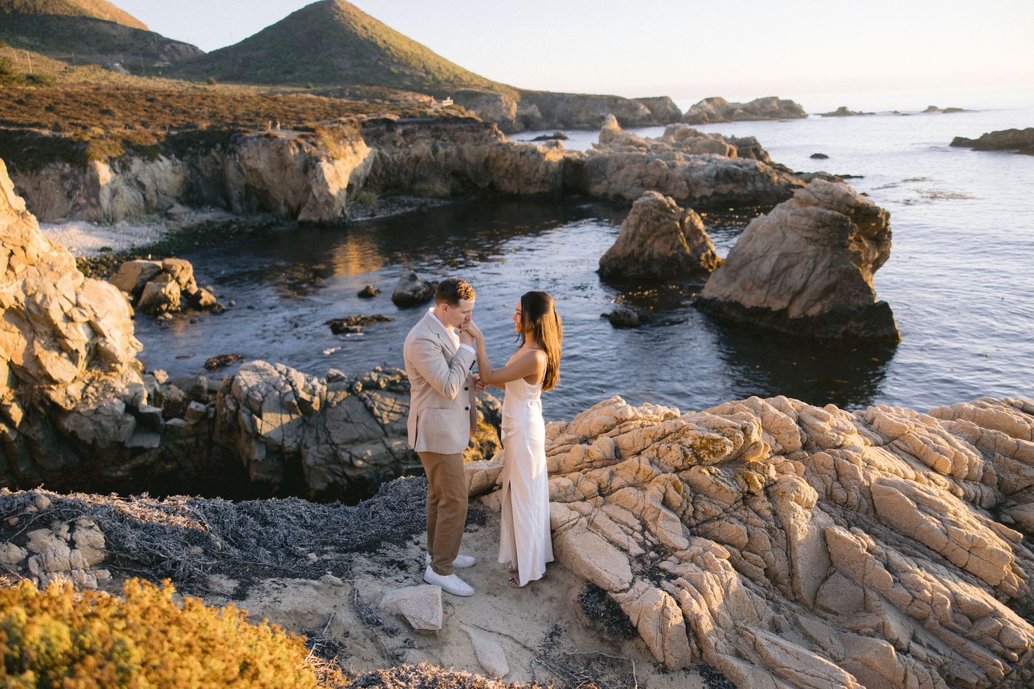 Two people standing on rocky coastal cliffs at sunset with waves crashing against the Big Sur shoreline in California.