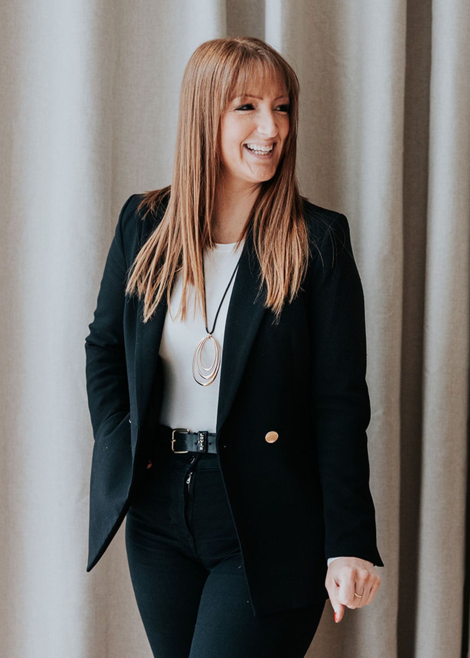 Professional woman in black blazer and white top smiles warmly while wearing long pendant necklace against neutral background.