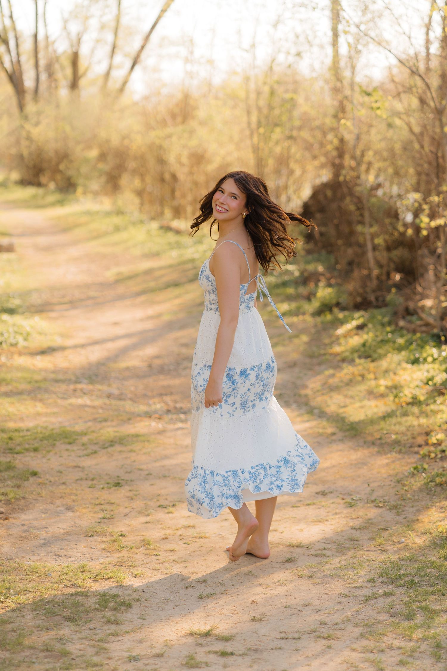 A person in a white and blue floral dress dances and twirls on a sunlit dirt path surrounded by trees.