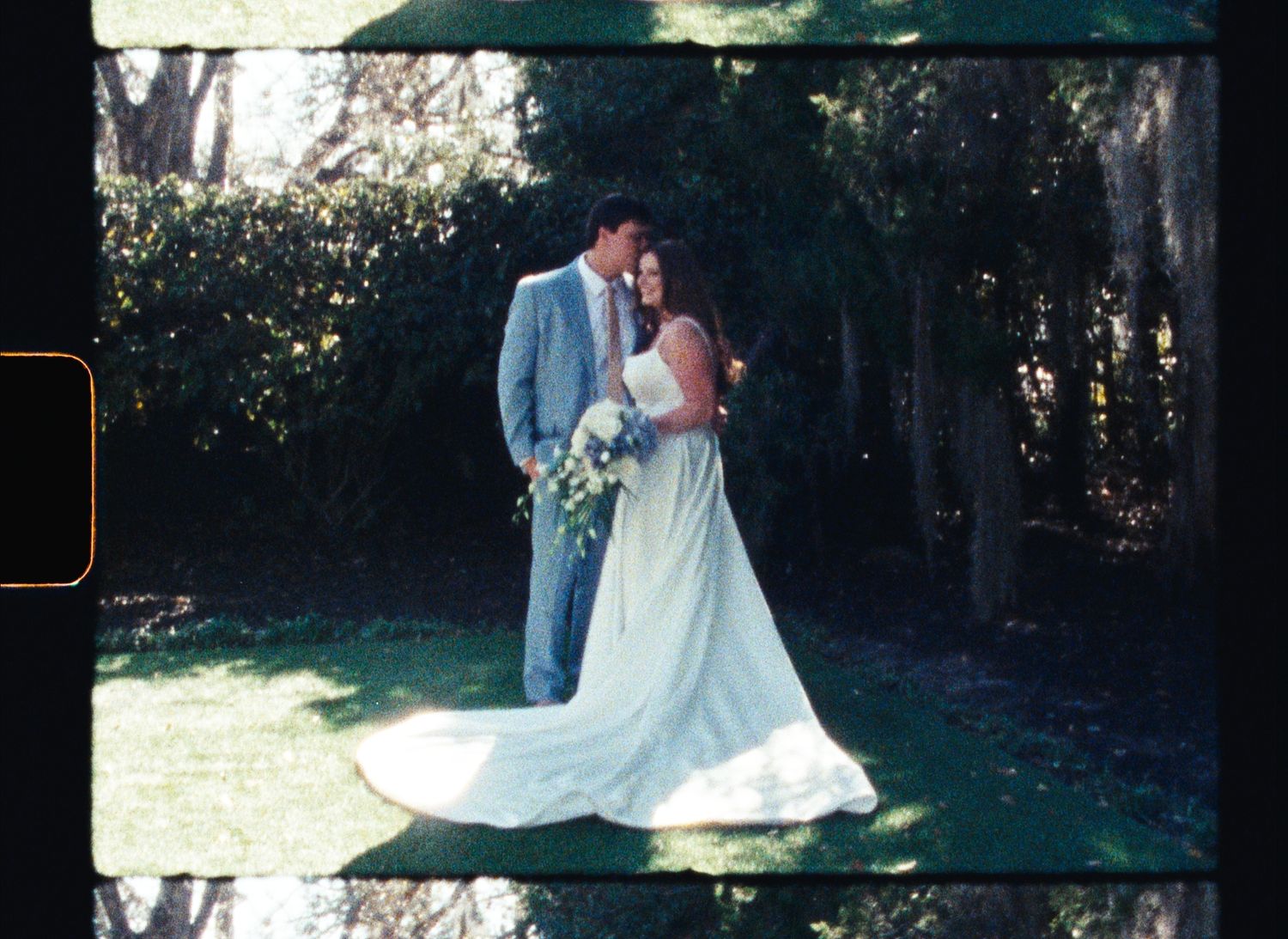 A couple embraces outdoors in a garden setting with the bride wearing a flowing white gown and holding a floral bouquet.