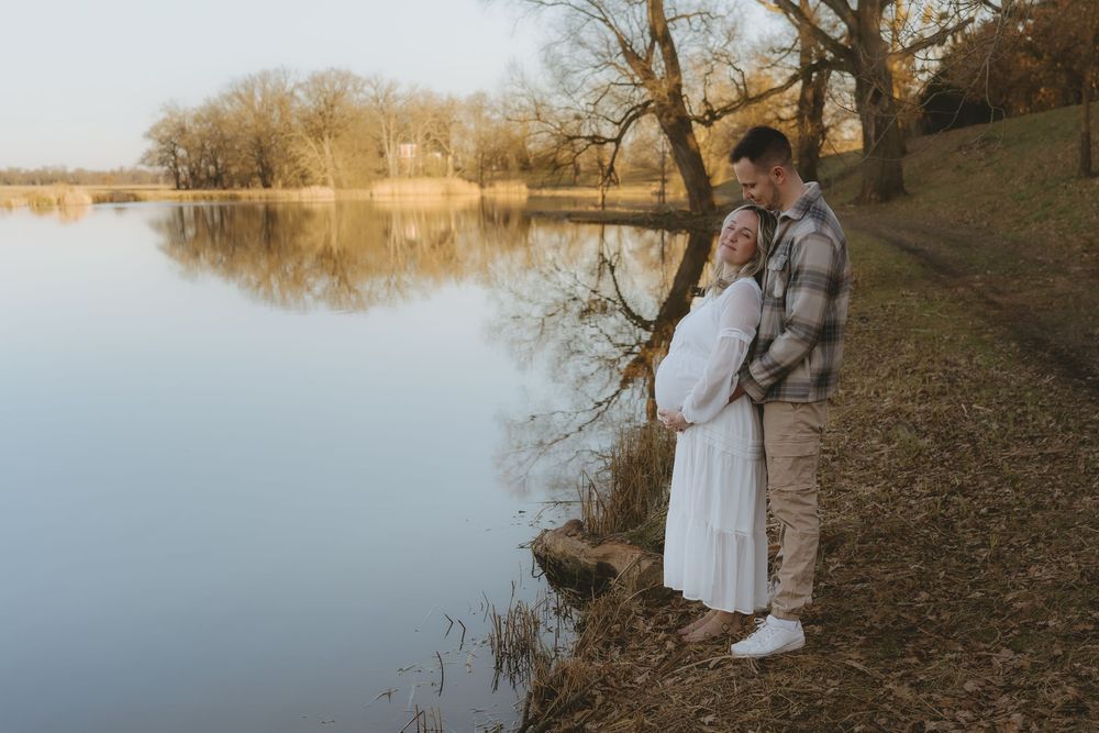 A couple stands together on a serene lakeside path during golden hour, surrounded by autumn trees and calm waters.