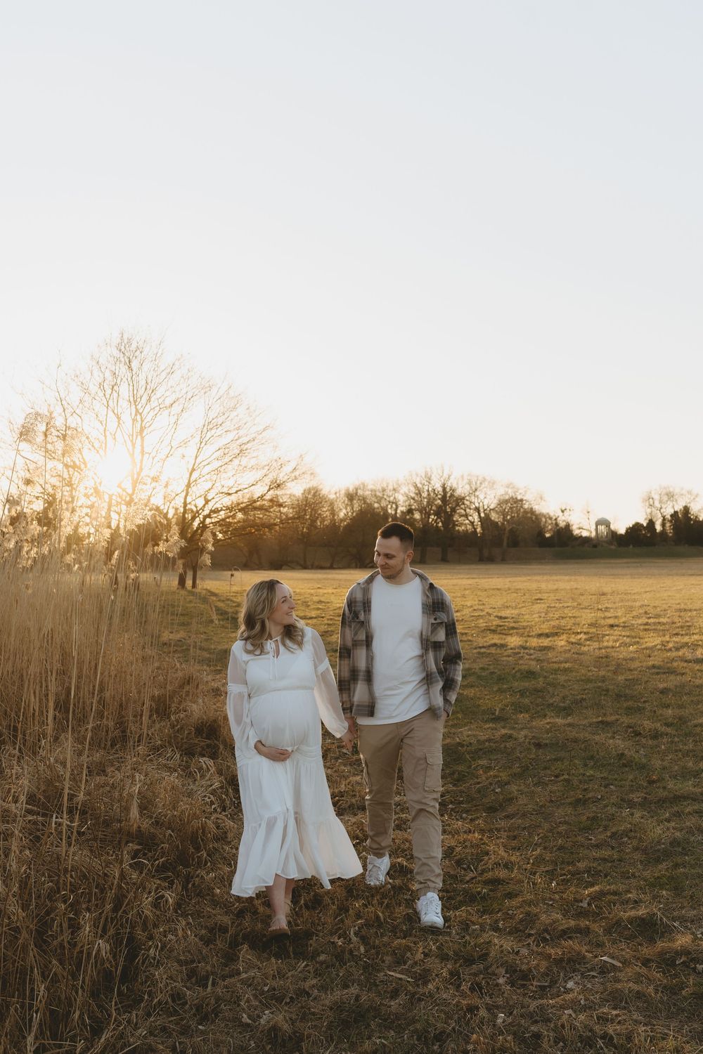 Expectant couple walking together in a golden field at sunset with tall grass and bare trees in background.