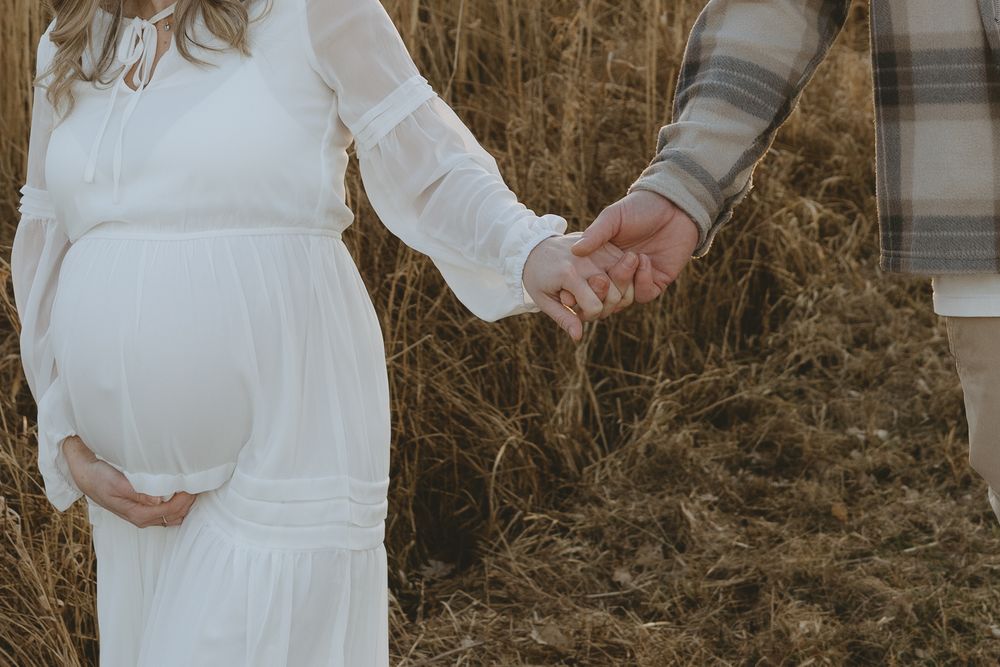 A pregnant woman in a white flowy dress holds hands with someone in a plaid shirt against dried tall grass.