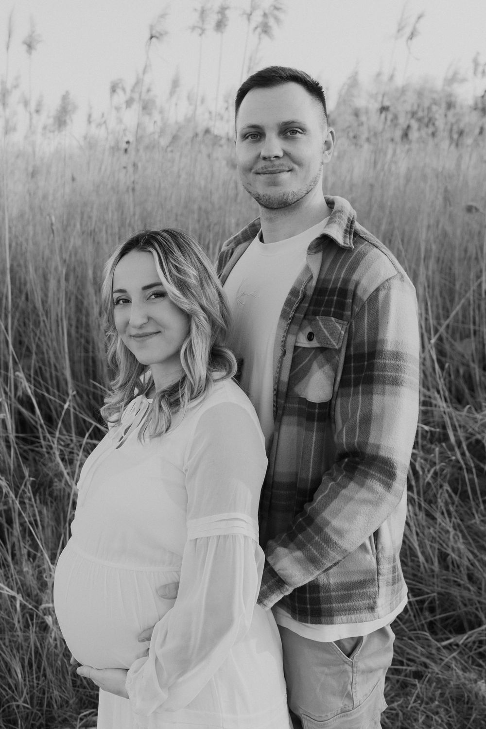 A couple stands together in a field of tall grass, with her wearing a white dress and him in a plaid shirt.